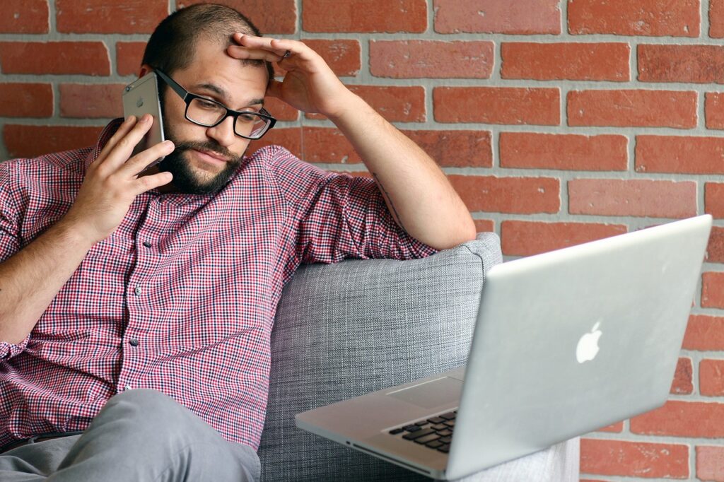 Man with a beard, on the phone looking at a laptop and feeling very frustrated.